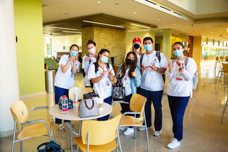 A group of nursing students wearing scrubs and masks stand next to a table and chairs in Cooper Hall, making the W sign with their hands.