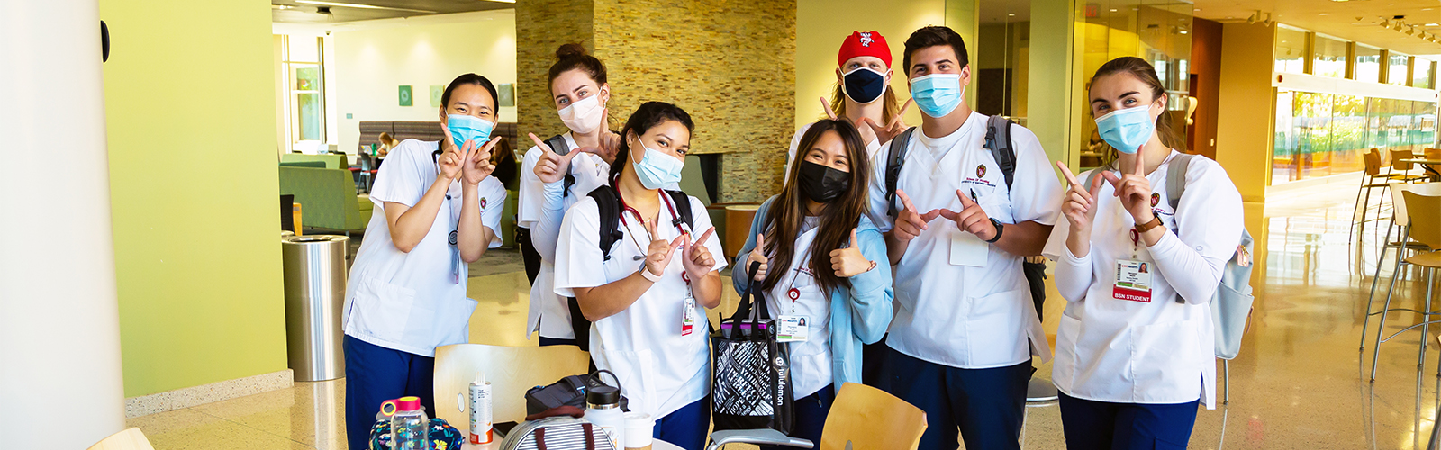 A group of nursing students wearing scrubs and masks stand next to a table and chairs in Cooper Hall, making the W sign with their hands.